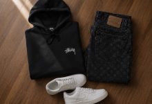 Black Stussy hoodie folded next to dark monogram denim and chunky sneakers on a wooden floor