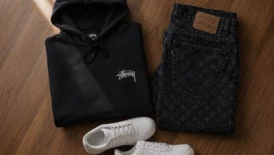 Black Stussy hoodie folded next to dark monogram denim and chunky sneakers on a wooden floor